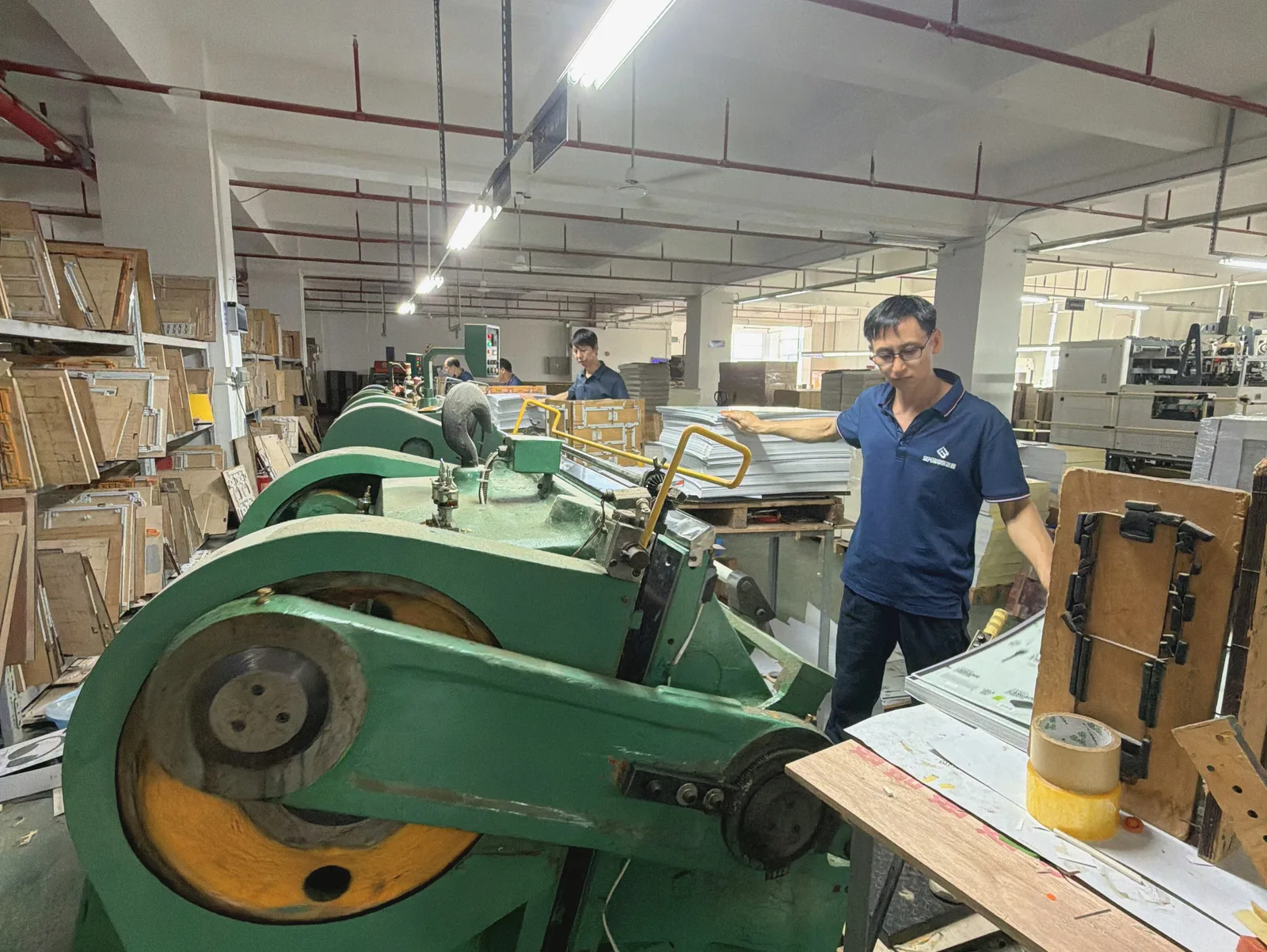 Operator beside die-cutting equipment and stacked board sheets in a packaging workshop